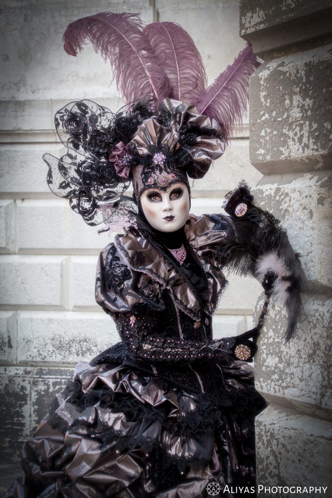 On this picture, you can see a woman wearing a silver-lilac-black costume in the Carnival of Venice 2019. She wears an elaborate head piece with black lace and lilac feathers.
