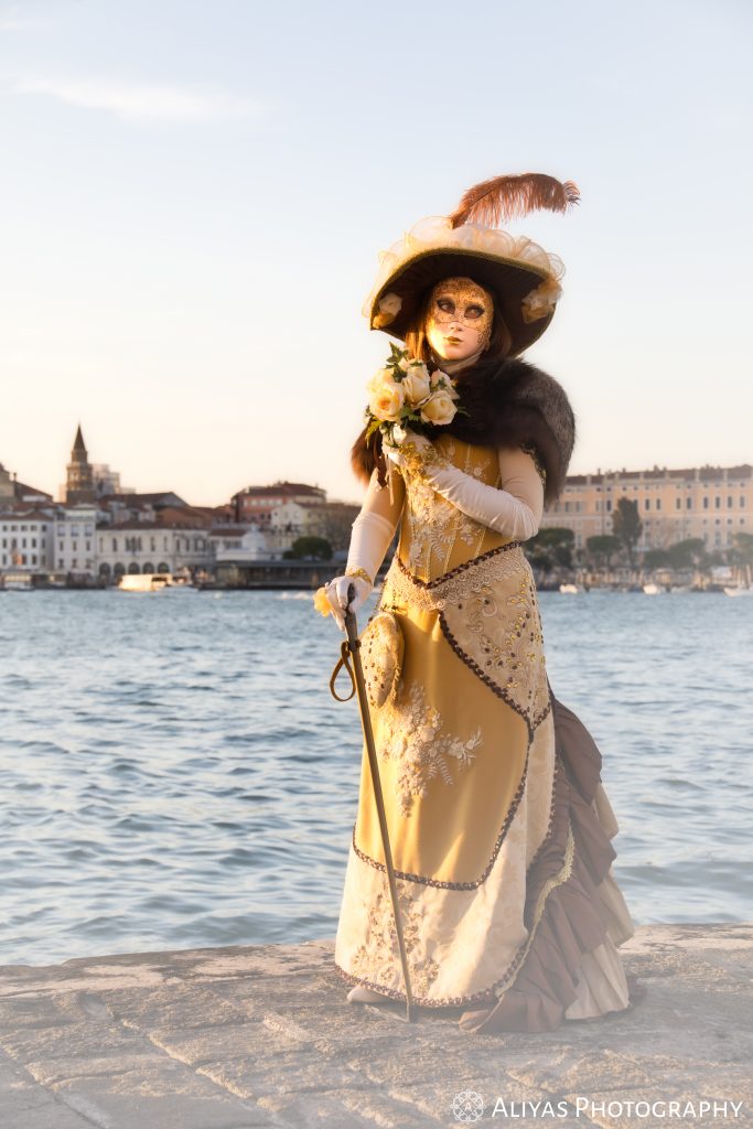On this picture, you can see a woman wearing a golden white costume in the Carnival of Venice in 2020. Her accessory is a bunch of pale yellow roses and a walking stick.