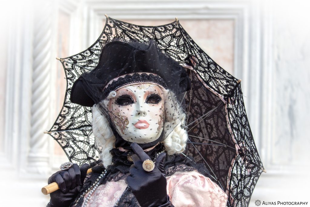 On this picture, you can see a woman wearing a black-pink costume in the Carnival of Venice 2018. Her accessories are a black and pink lace umbrella and a golden ring.